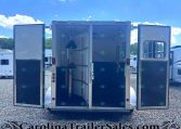 Open horse trailer with doors wide, showing empty interior stalls and equipment inside, on a gravel lot under a bright blue sky with clouds, watermark at bottom