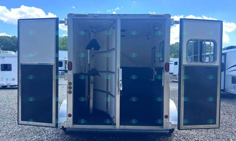Open horse trailer with doors wide, showing empty interior stalls and equipment inside, on a gravel lot under a bright blue sky with clouds, watermark at bottom