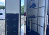 Open horse trailer with individual stalls and saddle racks, doors ajar in a gravel lot.