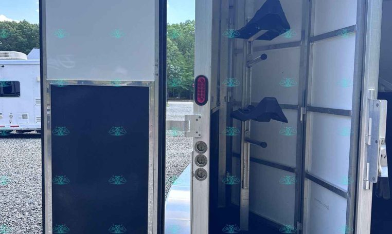 Open horse trailer with individual stalls and saddle racks, doors ajar in a gravel lot.