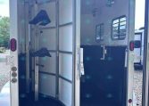 Interior view of an open horse trailer with divided stalls, hanging hooks, and a small blue saddle rack; rear doors open onto gravel ground.