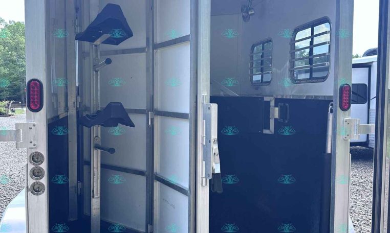 Interior view of an open horse trailer with divided stalls, hanging hooks, and a small blue saddle rack; rear doors open onto gravel ground.