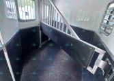 Interior of a horse trailer: aluminum divider/rail, black lower wall panels, and a speckled non-slip floor; windows with bars on the sides and a small hatch/door at the back.