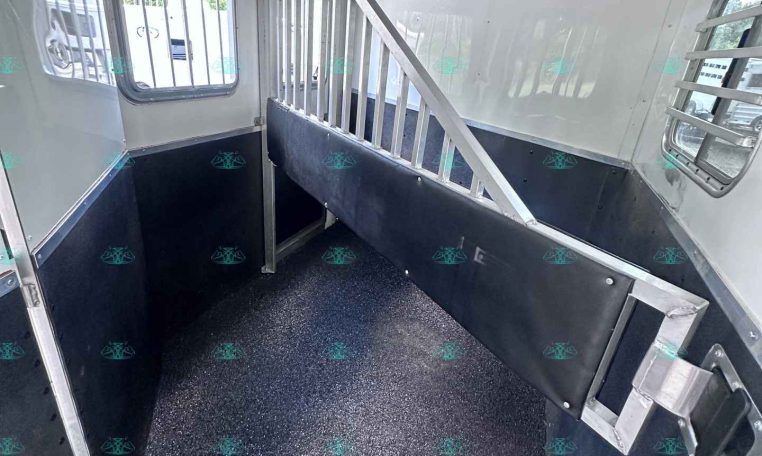 Interior of a horse trailer: aluminum divider/rail, black lower wall panels, and a speckled non-slip floor; windows with bars on the sides and a small hatch/door at the back.