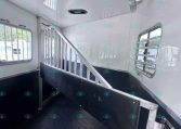 Interior of a white horse trailer showing a metal stall divider, raised walkway, and ventilation windows with bars; black rubberized lower walls and a mounted fan on the wall.