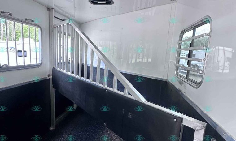 Interior of a white horse trailer showing a metal stall divider, raised walkway, and ventilation windows with bars; black rubberized lower walls and a mounted fan on the wall.