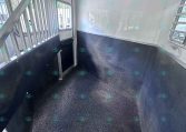 Interior of a white horse trailer: narrow space with a metal divider, grated door, and windows with bars along the left wall.