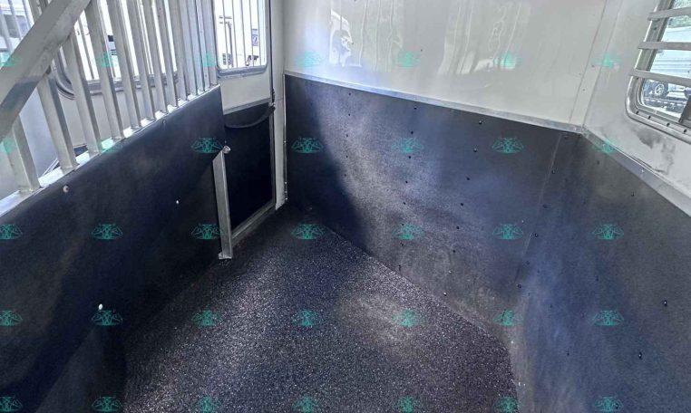 Interior of a white horse trailer: narrow space with a metal divider, grated door, and windows with bars along the left wall.