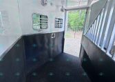 Interior of a white horse trailer viewed from inside, showing black lower walls, small barred windows, ceiling fans, and a grated doorway to an exterior ramp.