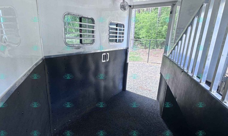 Interior of a white horse trailer viewed from inside, showing black lower walls, small barred windows, ceiling fans, and a grated doorway to an exterior ramp.