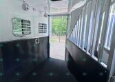 Interior of a horse trailer with metal stall dividers on the right, ventilated windows, ceiling fans, and a non-slip rubber floor.