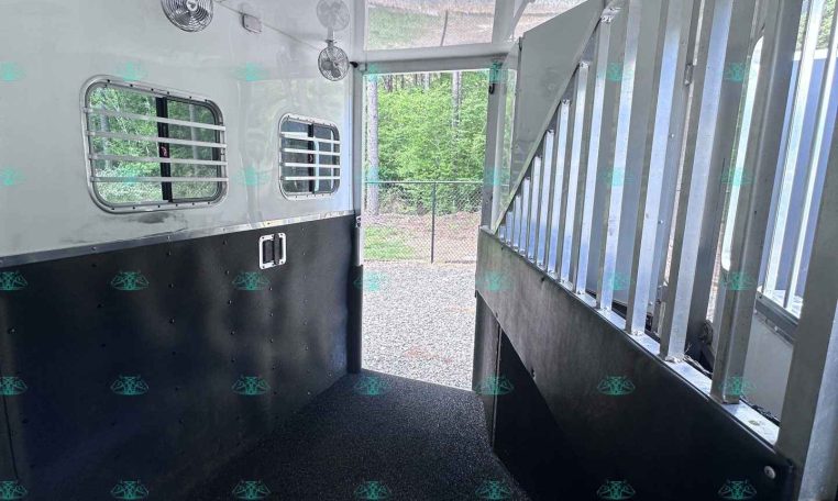 Interior of a horse trailer with metal stall dividers on the right, ventilated windows, ceiling fans, and a non-slip rubber floor.