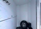 Interior of a white trailer cargo area with multiple black U-shaped hooks along the left wall and a mounted spare tire at the far end.