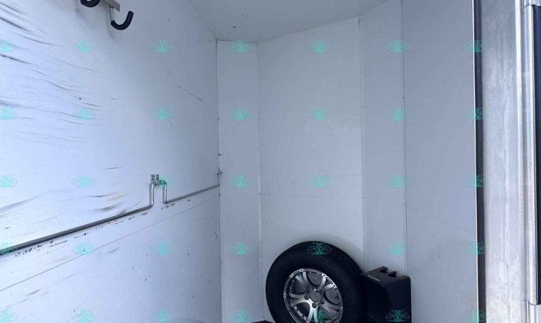 Interior of a white trailer cargo area with multiple black U-shaped hooks along the left wall and a mounted spare tire at the far end.