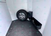 Inside a cargo trailer showing a mounted tire, spare wheel, and a black storage bin against light gray wall panels on a carpeted floor