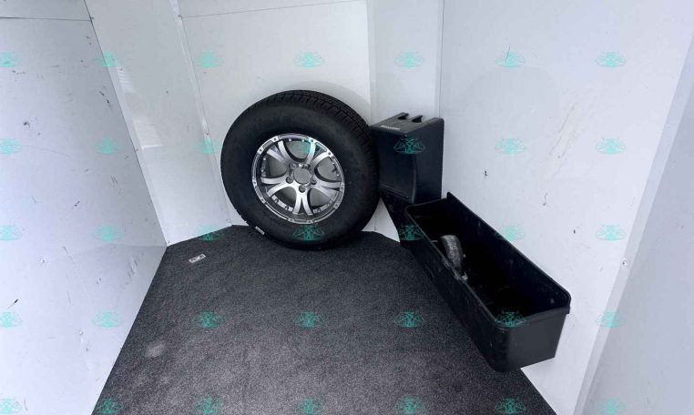 Inside a cargo trailer showing a mounted tire, spare wheel, and a black storage bin against light gray wall panels on a carpeted floor