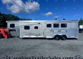 Silver Merhow horse trailer on a gravel lot with two windows, under a blue sky with clouds; red machinery on the left and a watermark bottom edge reading CarolinaTrailersales.com.