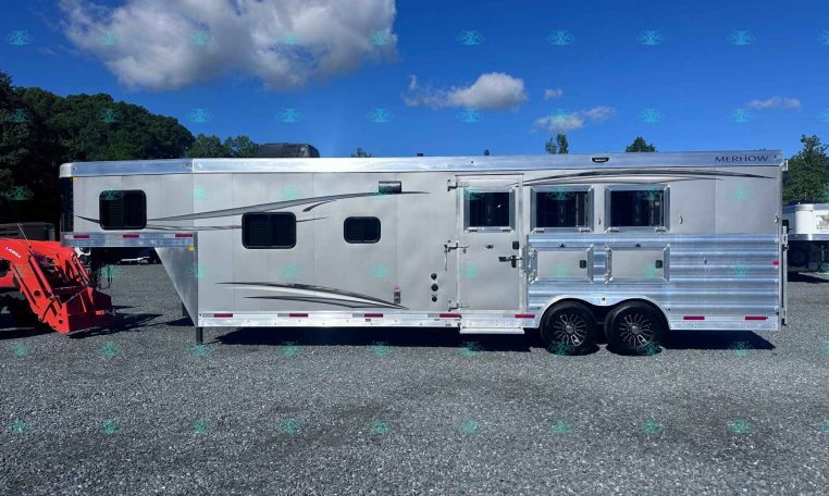 Silver Merhow horse trailer on a gravel lot with two windows, under a blue sky with clouds; red machinery on the left and a watermark bottom edge reading CarolinaTrailersales.com.