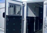 Open aluminum horse trailer with a barred vent window and interior doorway, parked on gravel under a blue sky.