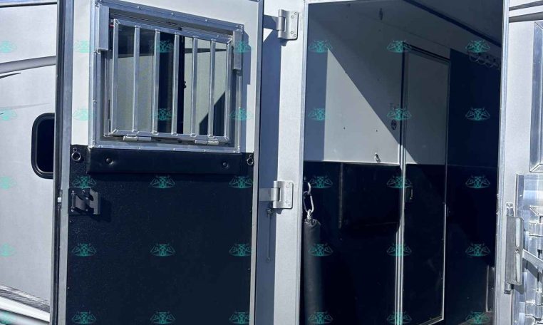 Open aluminum horse trailer with a barred vent window and interior doorway, parked on gravel under a blue sky.