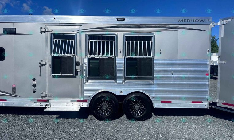 Silver horse trailer with three stall doors, barred windows, and chrome trim sits on gravel under a clear blue sky.