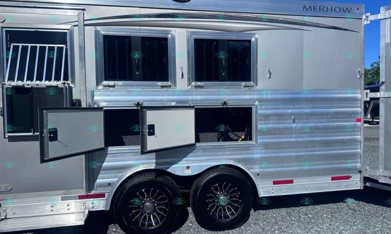 Side view of a shiny silver Merhow horse trailer with open storage compartments and windows behind a clear blue sky.