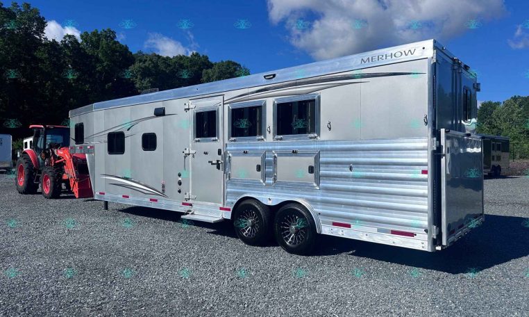 Silver Merhow horse trailer with multiple windows and side storage bays on a gravel lot under a blue sky.