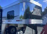 Shiny silver aluminum trailer with a mountain logo, parked on a gravel lot with propane tanks underneath.