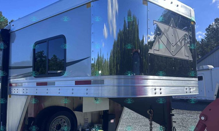 Shiny silver aluminum trailer with a mountain logo, parked on a gravel lot with propane tanks underneath.