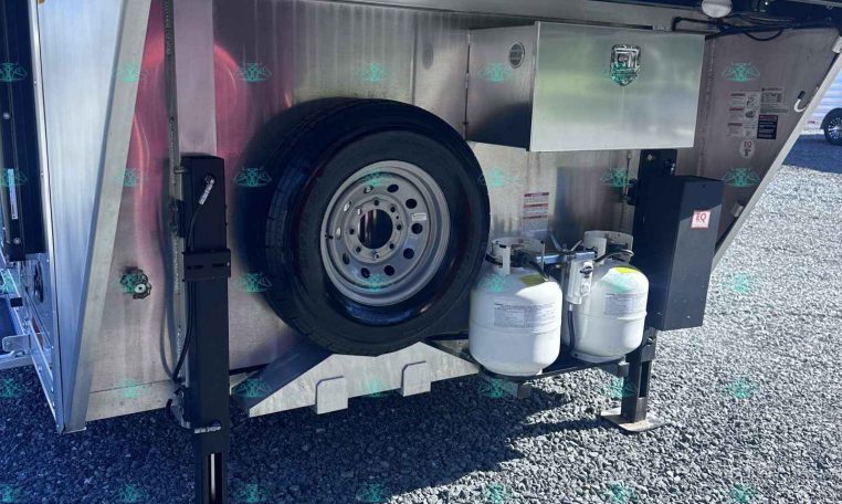 Open storage compartment of a utility trailer showing a mounted spare tire and propane tanks.