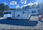 White fifth-wheel RV trailer parked on a gravel lot with tall pine trees and a clear blue sky behind it.