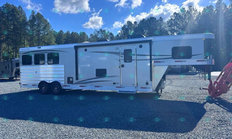 White fifth-wheel RV trailer parked on a gravel lot with tall pine trees and a clear blue sky behind it.