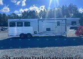 White Merhow horse trailer on a gravel lot with tall trees and a blue sky in the background; features multiple side windows and a side door, twin-axle trailer on display at a dealership lot.
