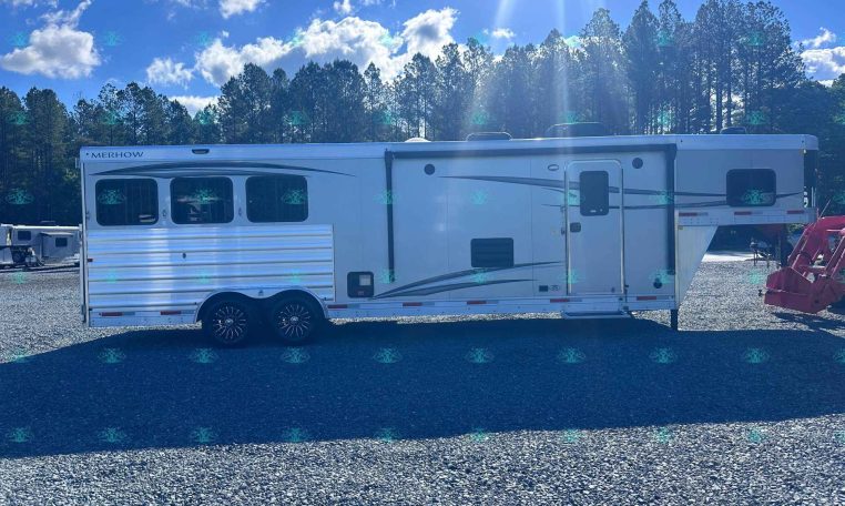 White Merhow horse trailer on a gravel lot with tall trees and a blue sky in the background; features multiple side windows and a side door, twin-axle trailer on display at a dealership lot.