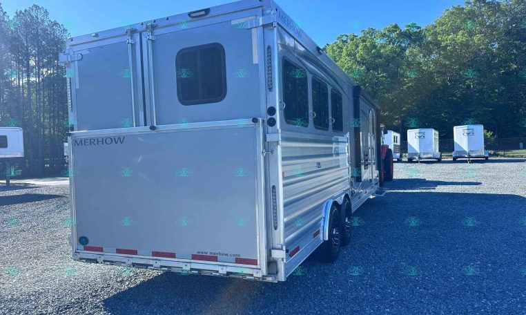Silver Merhow horse trailer parked on a gravel lot with trees in the background and other trailers in the distance under a clear blue sky