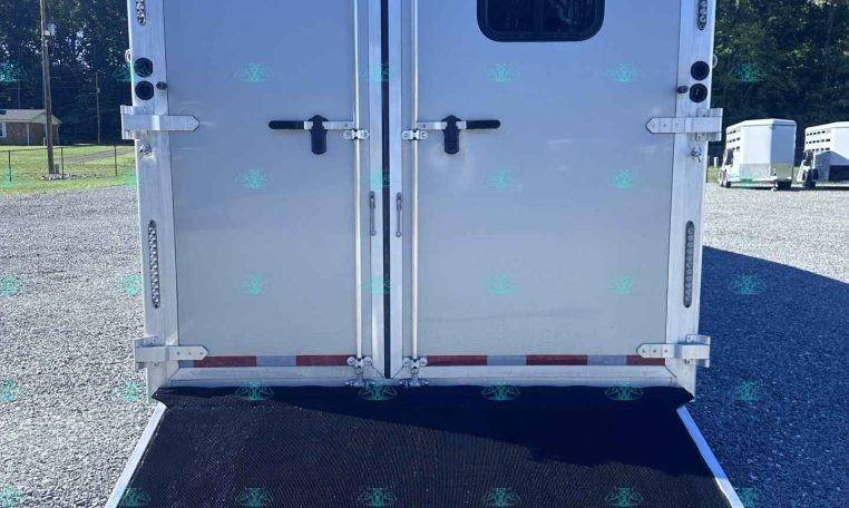 Rear of a silver cargo trailer with double doors, a small window on the right door, and a lowered loading ramp on a gravel lot.
