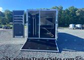 Open horse trailer with stalls and saddle racks, parked on a gravel lot with other trailers in the background.