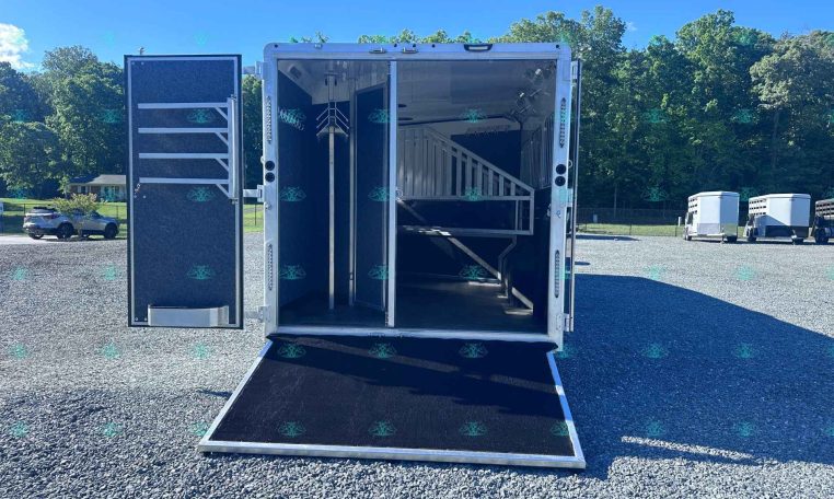 Open horse trailer with stalls and saddle racks, parked on a gravel lot with other trailers in the background.