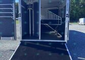 Horse trailer with its rear ramp lowered, revealing interior stalls and a staircase in an outdoor lot under a clear blue sky.
