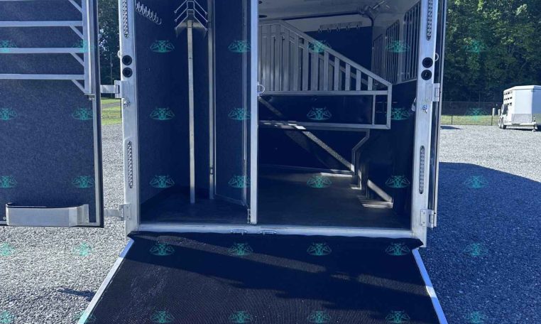 Horse trailer with its rear ramp lowered, revealing interior stalls and a staircase in an outdoor lot under a clear blue sky.