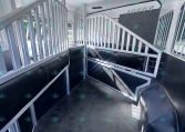 Interior of a silver aluminum trailer with partitioned stalls and metal railings, black rubber flooring, and overhead vents.