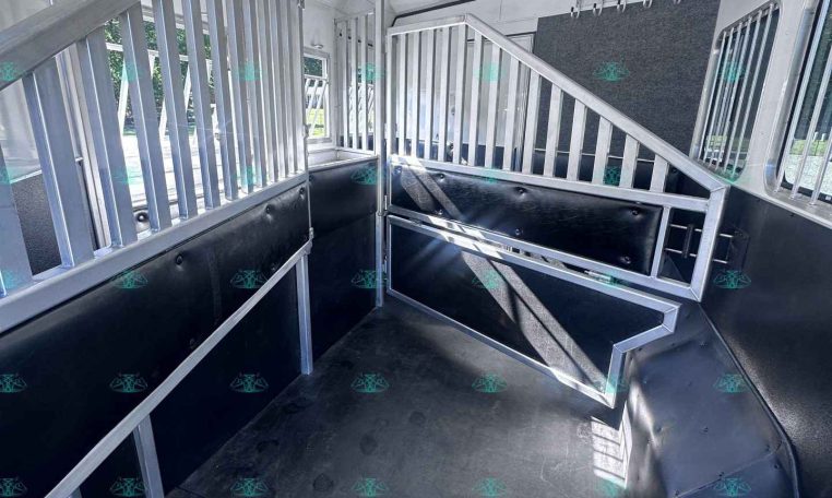 Interior of a silver aluminum trailer with partitioned stalls and metal railings, black rubber flooring, and overhead vents.