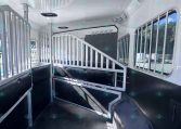 Interior of a white horse trailer with partitioned stalls, metal rails, and a ramp area visible near the bottom left.