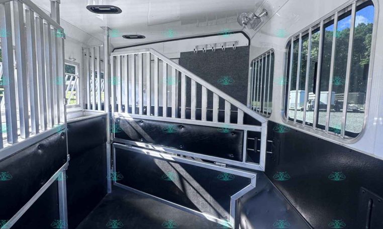 Interior of a white horse trailer with partitioned stalls, metal rails, and a ramp area visible near the bottom left.