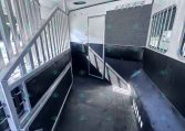 Interior of a horse trailer showing metal stalls, black rubber flooring, and a rear door with windows on the right side wall.