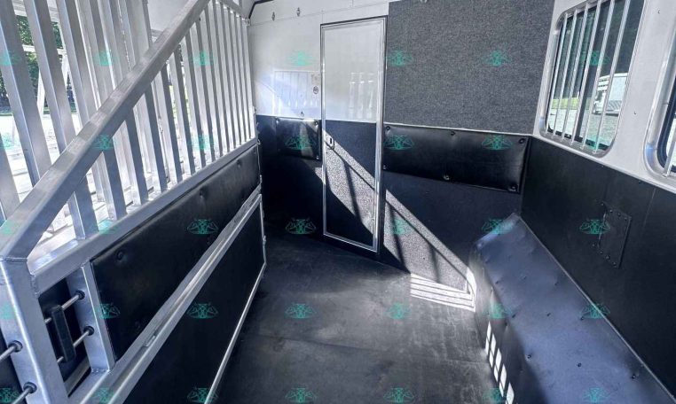 Interior of a horse trailer showing metal stalls, black rubber flooring, and a rear door with windows on the right side wall.