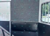 Interior of a trailer showing a grey carpeted divider with five metal hooks mounted along the top, black padded lower wall, and a window with bars to the right.