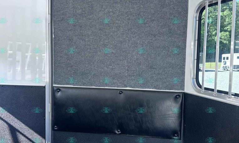 Interior of a trailer showing a grey carpeted divider with five metal hooks mounted along the top, black padded lower wall, and a window with bars to the right.