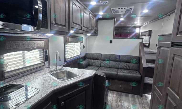 Interior of a modern RV kitchen and living area with dark cabinetry, marble-like counters, stainless steel appliances, and a gray sofa.