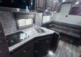 Interior of a modern RV kitchen with a marble-like countertop, stainless microwave, and dark wood cabinets above a sink and stove, next to a black leather sofa on the right (bright overhead lights).